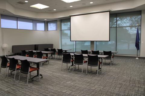 Photograph of meeting room with several rows of tables facing a large projector screen.