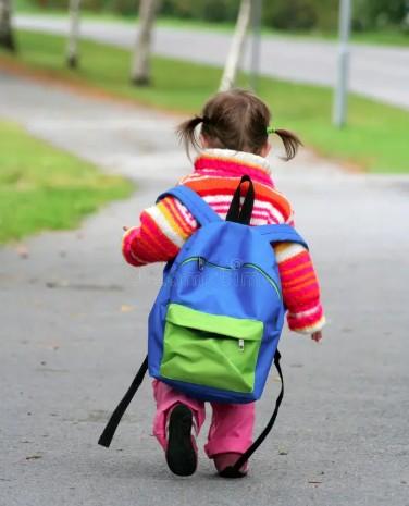 A young child with pigtails walks away down a sidewalk wearing a colorful striped sweater, pink pants, and a large blue and green backpack.
