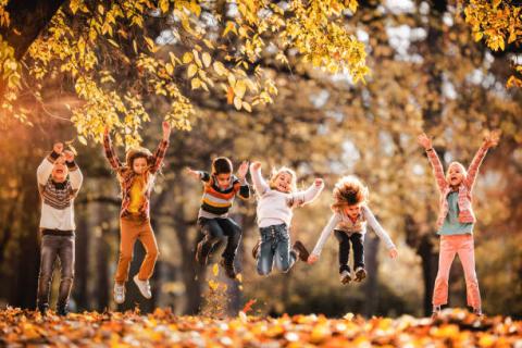 Group of six children smiling, jumping in the air, and playing in autumn leaves.