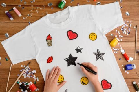 hand drawing designs on a white t-shirt laying on a wooden table surrounded by scattered art supplies.