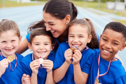 Group of smiling children wearing blue shirts and medals, standing together with an adult.
