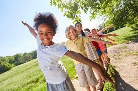 group of smiling children balanced with arms outstretched while walking in a line outdoors on a sunny day.