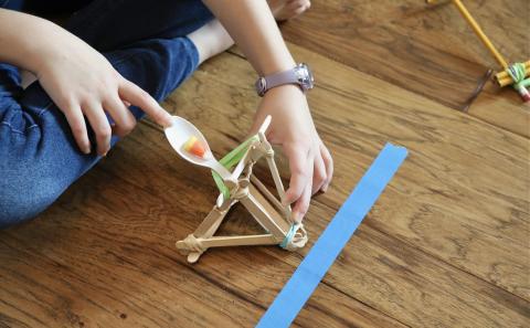 a child tests a homemade catapult made from craft sticks, rubber bands, and a plastic spoon holding a piece of candy corn on the floor.