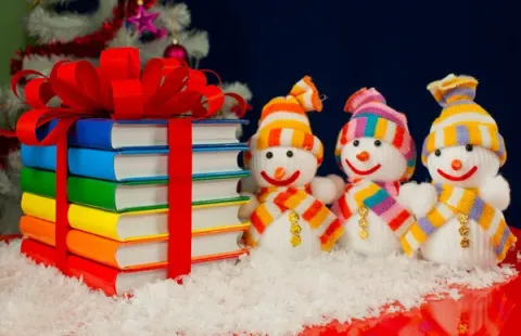 Three cheerful snowmen in colorful hats and scarves stand beside a stack of rainbow-colored books tied with a red ribbon, surrounded by artificial snow and holiday decor