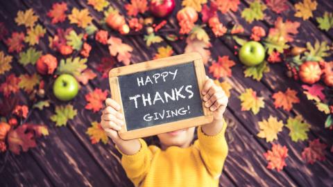 a child in a yellow sweater holds up a small chalkboard that reads "Happy Thanksgiving!" against a backdrop of autumn leaves and apples.