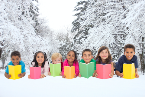 a group of smiling children hold colorful books while sitting in the snow, surrounded by snow-covered trees. 